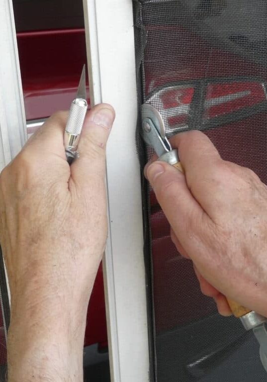 A homeowner is using a spline roller tool and knife to replace a damaged panel in a sliding garage screen door with a metal frame.